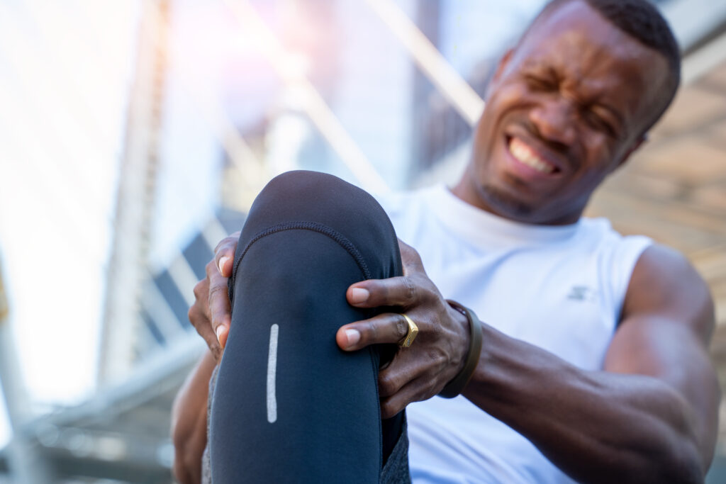 African american male runner bends over clutching his knee while in intense pain from an acute knee injury, athletes hurt the knees.