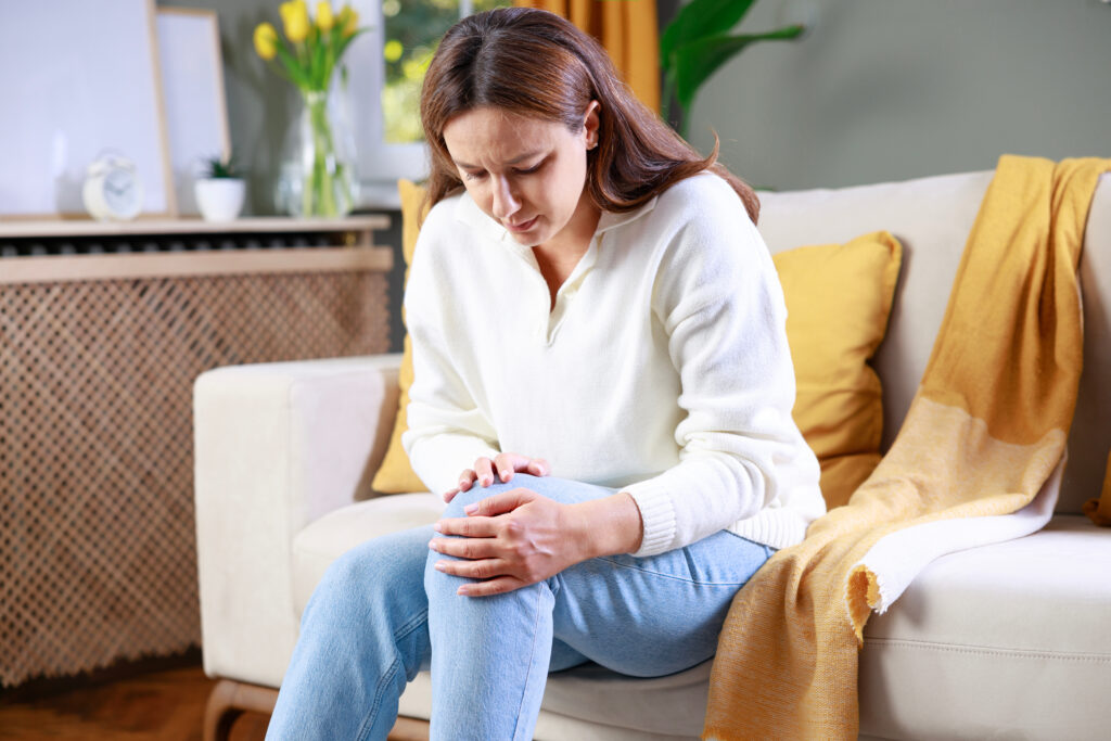 Young woman suffering from pain in knee. Mid adult age woman feeling unwell, suffering from pain in knee while sitting on sofa in the living room at home